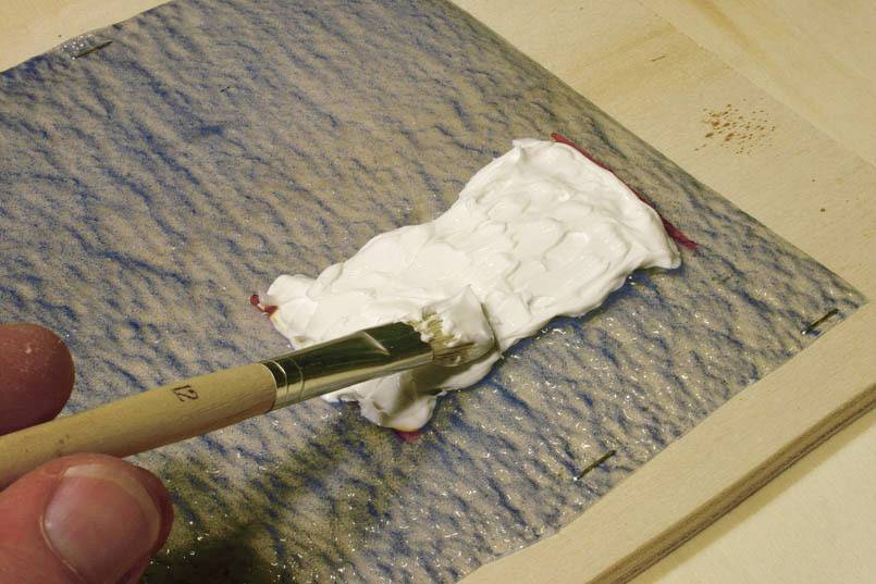 Een kwast verspreidt witte kleefpasta op een gestructureerd, blauw-beige oppervlak. De hand links houdt de kwast vast.