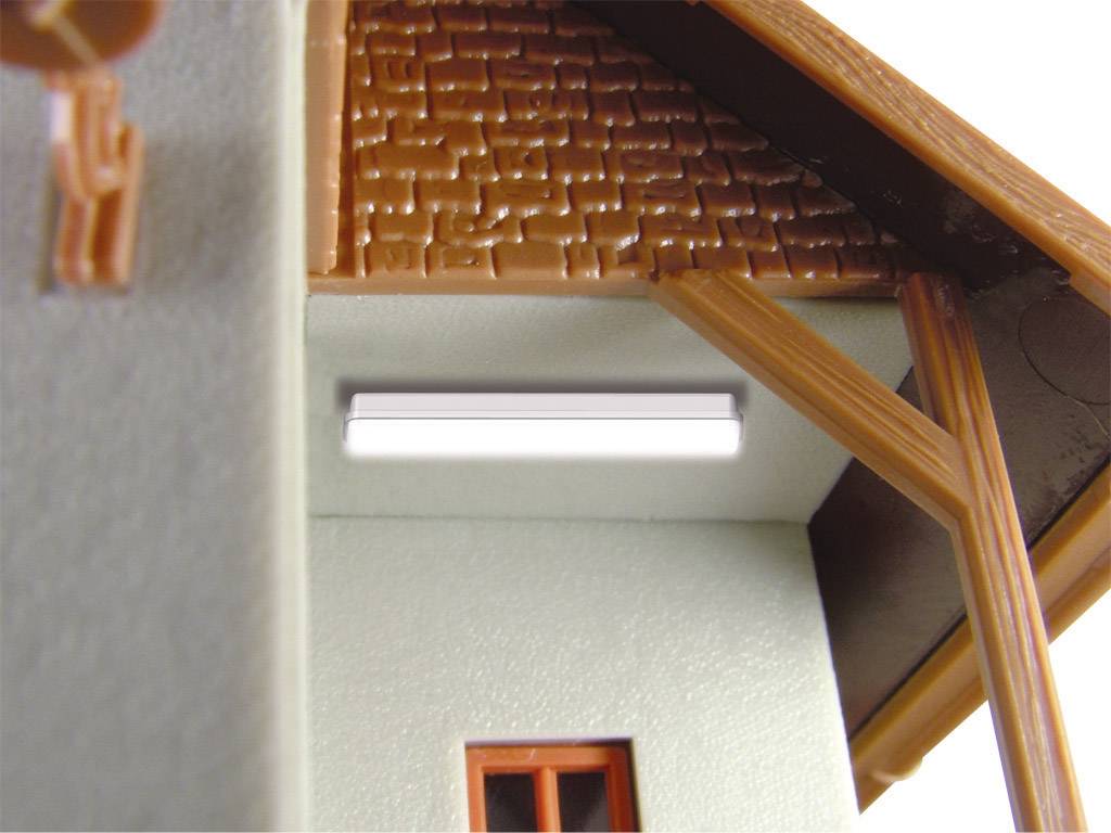 Deel van een miniatuurmodel van een huis met een zichtbaar dak en vensterramen, verlicht door een kleine, rechthoekige lamp.