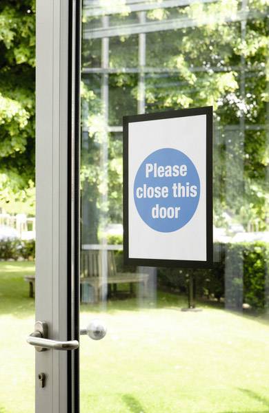 Een bordje aan een glazen deur met de tekst 'Gelieve deze deur te sluiten'. Op de achtergrond is een groene tuin zichtbaar.