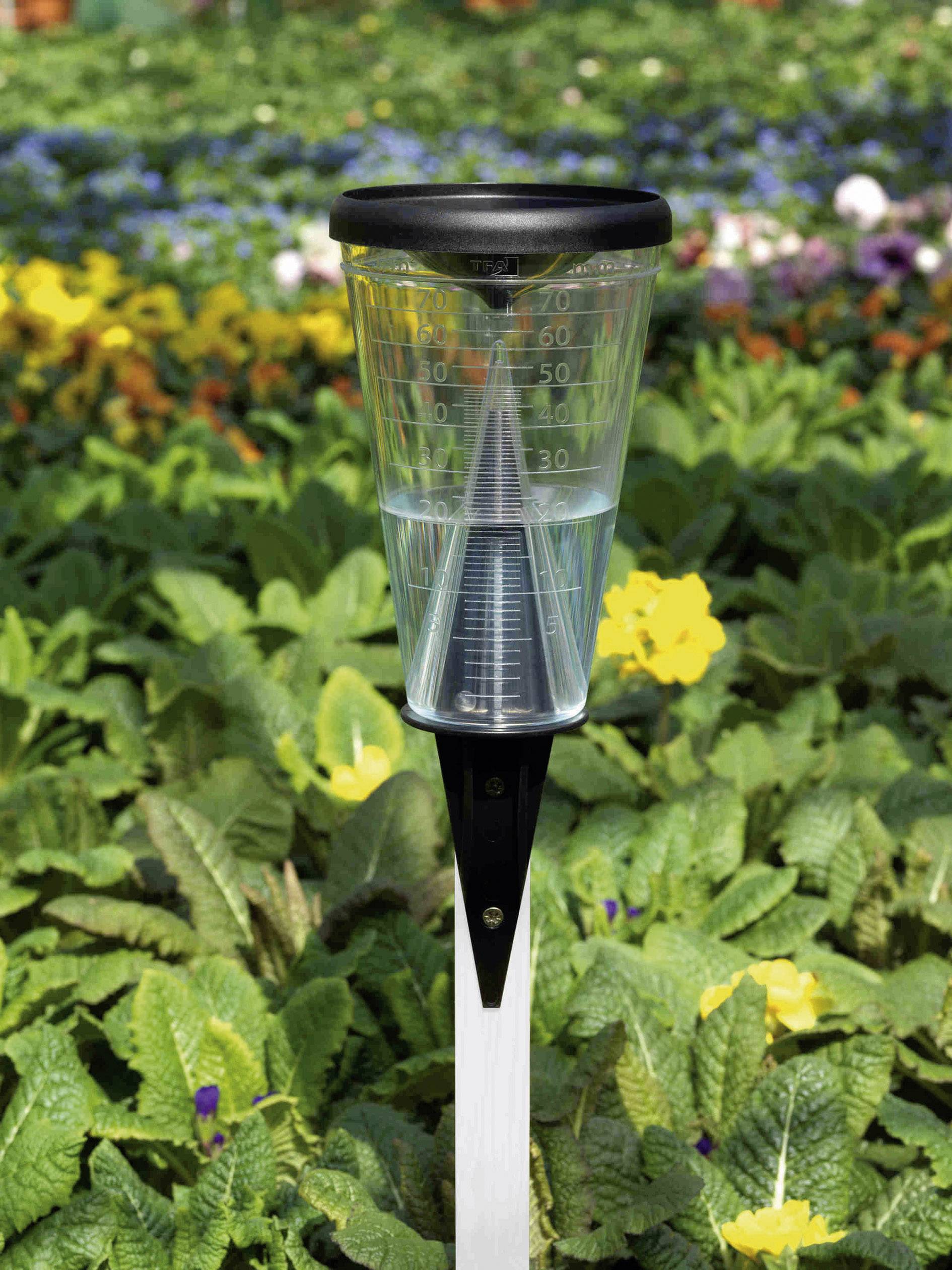 Regenmeter in de tuin, halfvol met water, omgeven door bloeiende planten op de achtergrond.