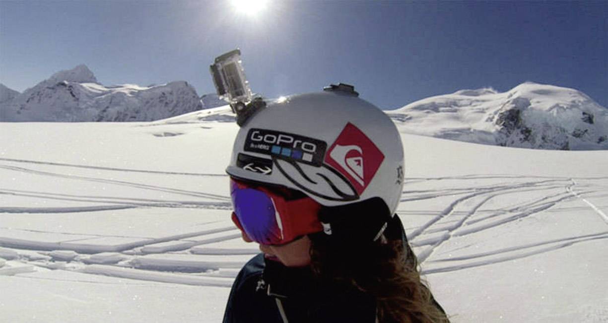 Een persoon met een helm en skibril rijdt in de zonneschijn op een besneeuwde berg. Een camera is aan de helm bevestigd.
