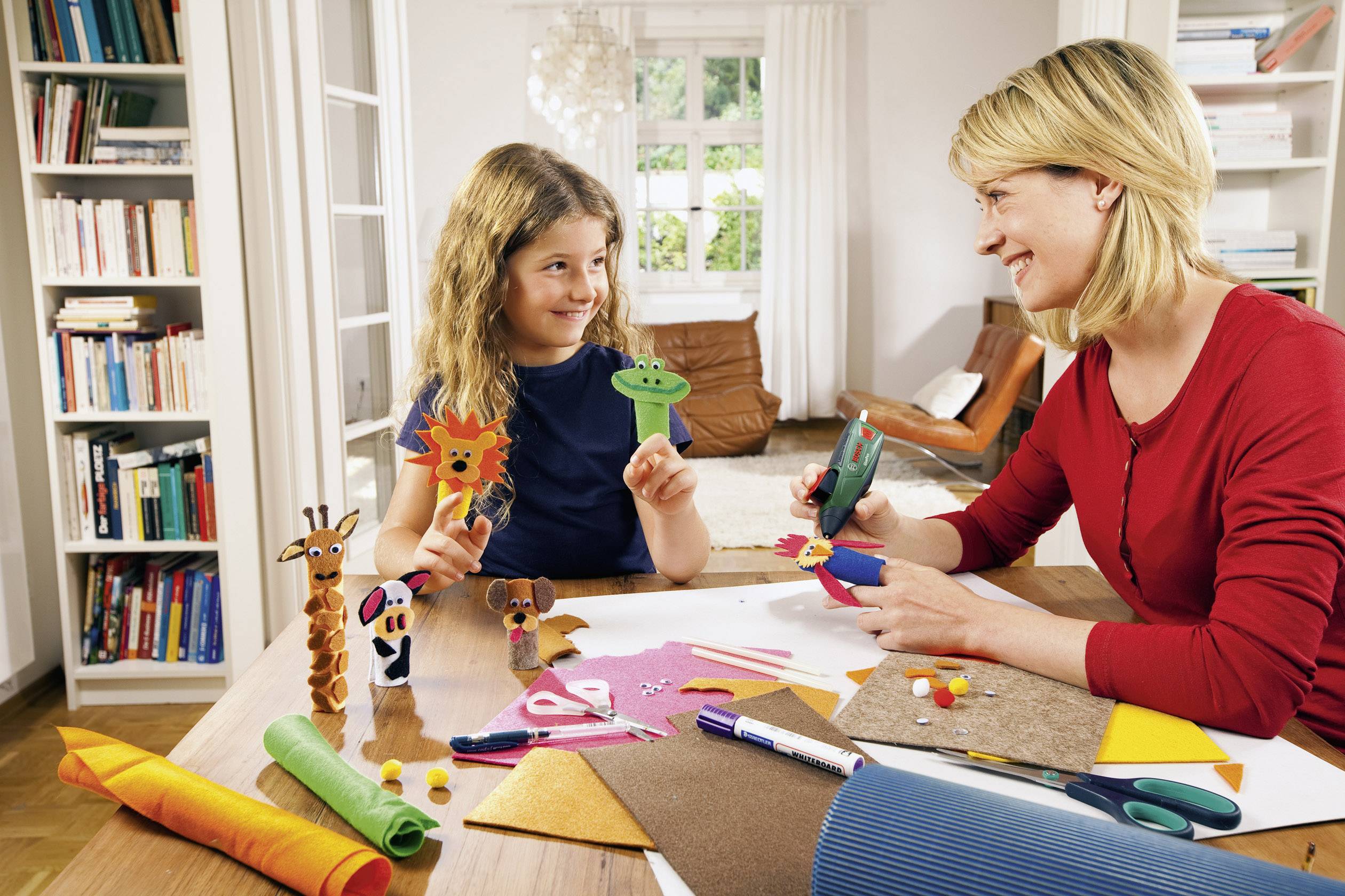 Een vrouw en een meisje knutselen figuurtjes van kleurrijk vilt aan een tafel vol knutselmateriaal in een gezellige ruimte.