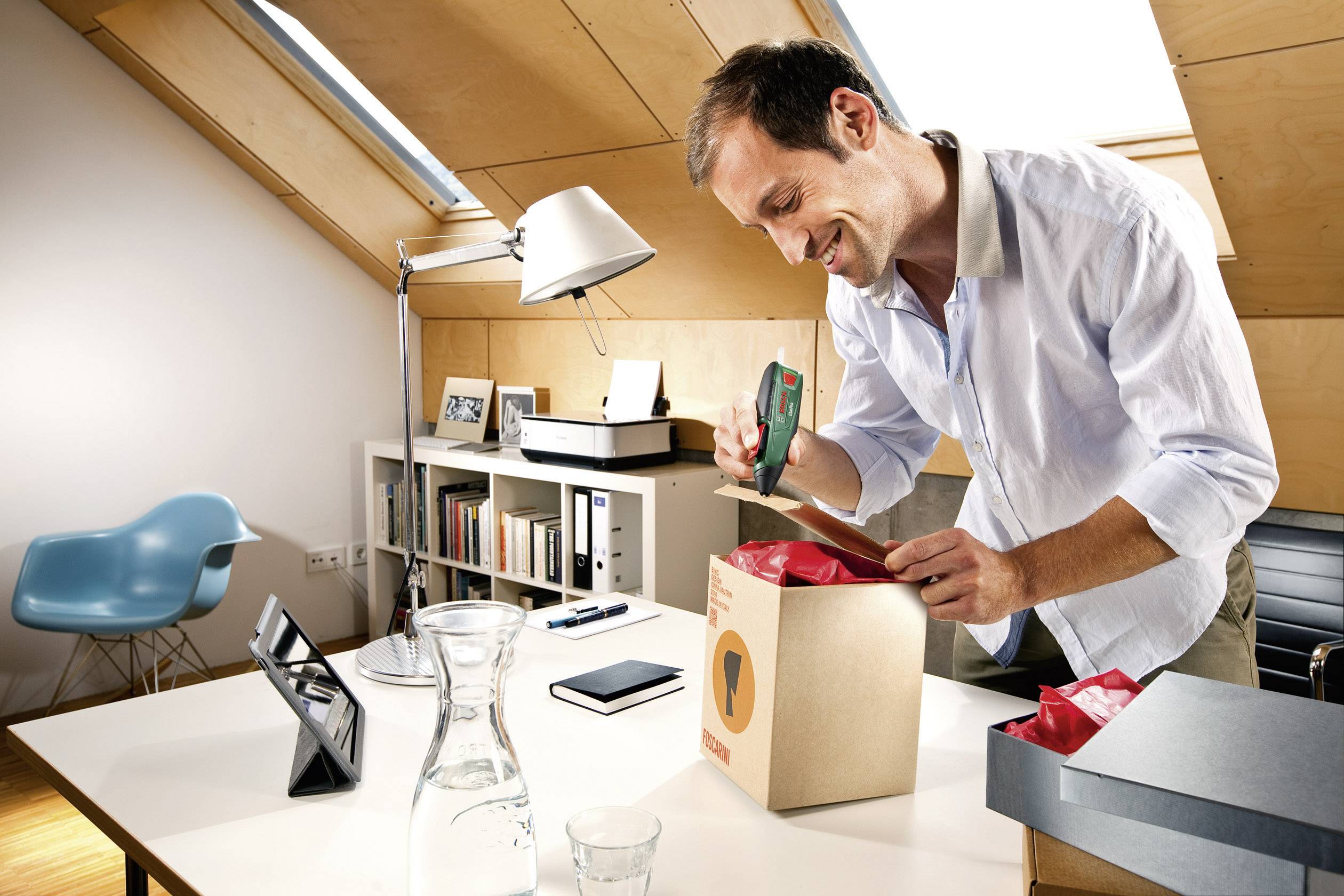 Een man glimlacht terwijl hij een pakket opent aan een bureau in een kantoor. De omgeving ziet er modern en licht uit.