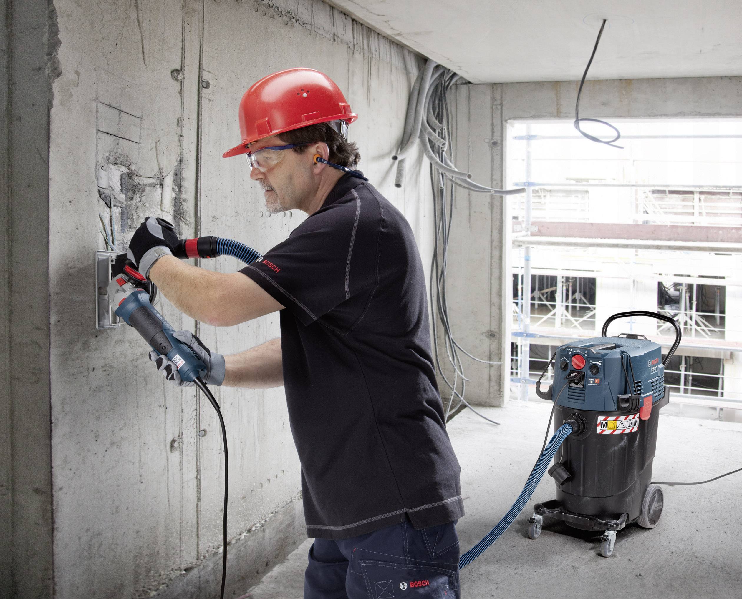 Een bouwvakker draagt een rode veiligheidshelm en gebruikt een boormachine aan een betonnen muur. Een industriële stofzuiger staat ernaast.