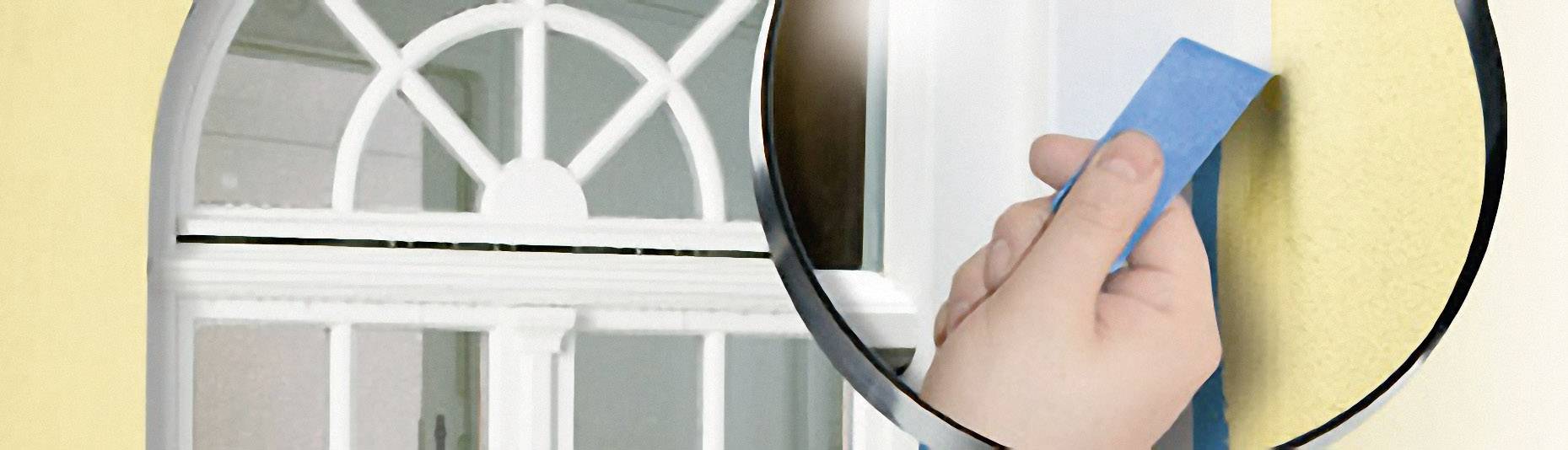 Een hand plakt blauwe schilderstape om het raam van een gele muur om het te beschermen tegen verfspetters.