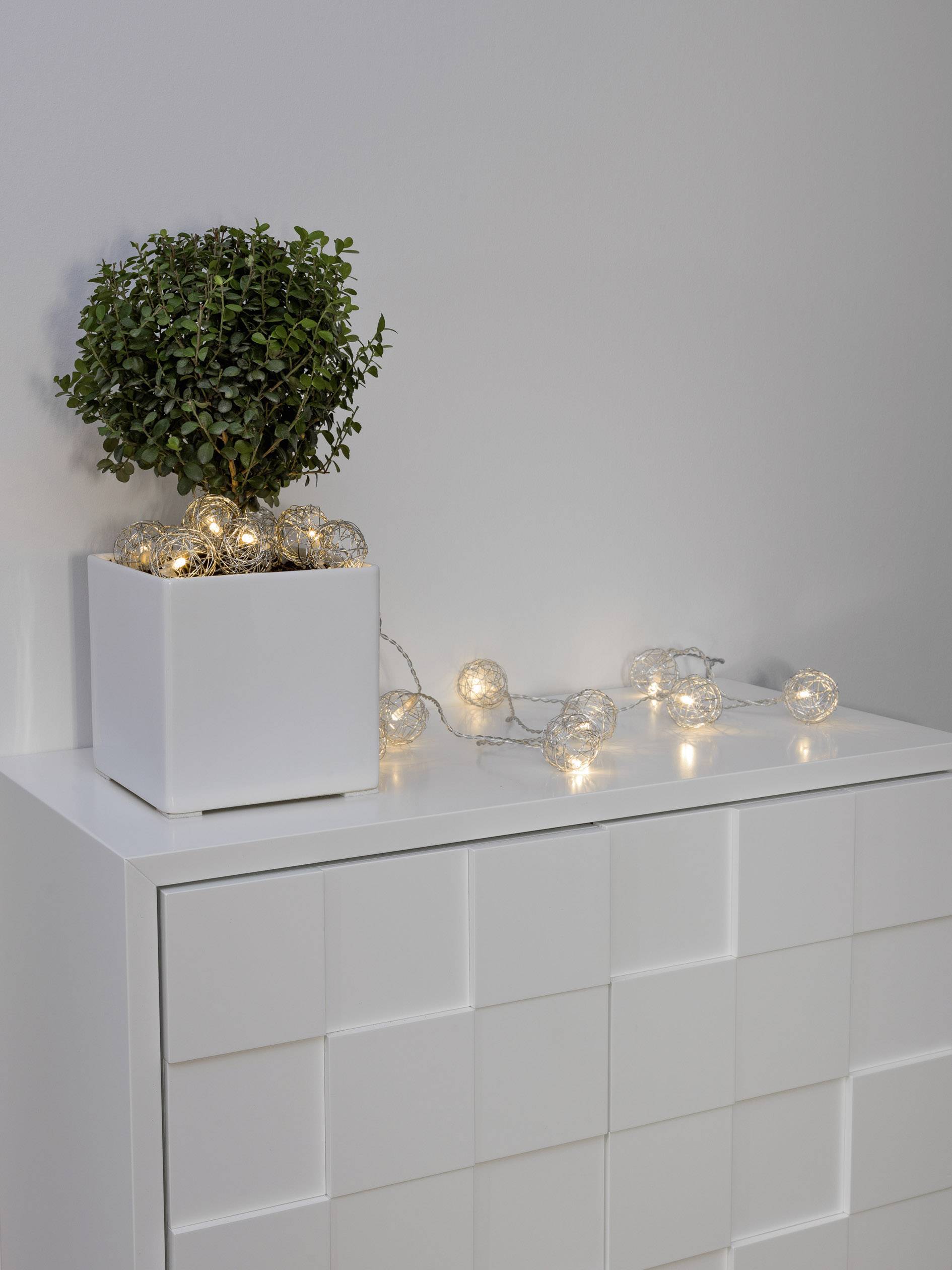 Witte ladenkast met geometrisch design, plant in witte pot en decoratieve lichtslingers bovenop. Strakke, moderne esthetiek.
