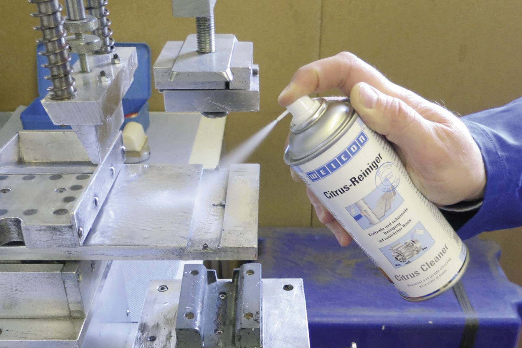 Een persoon spuit met een spray op een metalen machineoppervlak. De spray is geëtiketteerd als 'glasreiniger'.