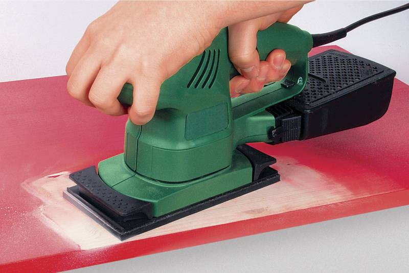 Een hand houdt een elektrische slijpmachine die wordt gebruikt op een rood gelakt houten bord om verf te schuren.