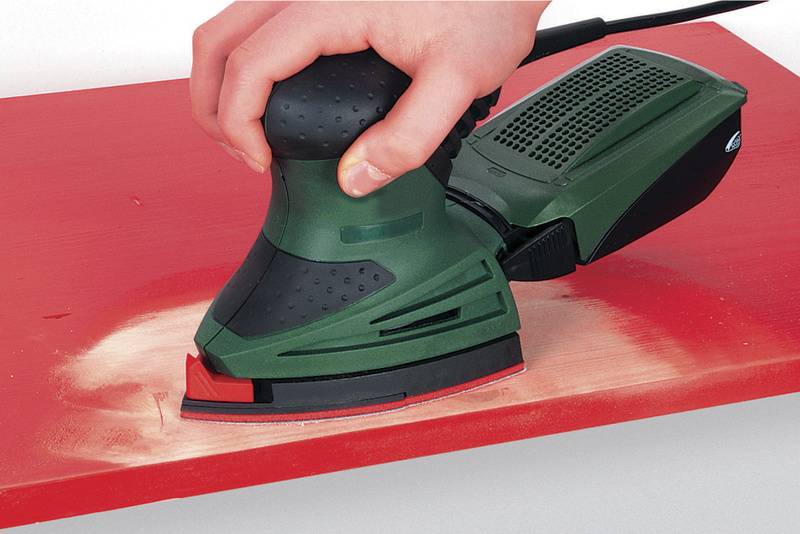 Een persoon slijpt met een groene elektrische slijpmachine een rode houten plaat, waarbij houtstof ontstaat.