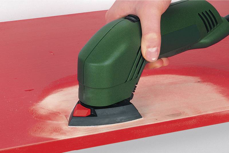 Een groene handmatige schuurmachine schuurt een rood houten oppervlak. Stof verzamelt zich tijdens het schuren.