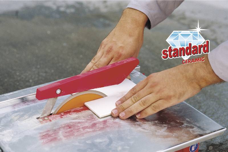 Een persoon snijdt een keramische tegel met een elektrische tegelsnijder. Tekst 'standaard CERAMIC' naast de handeling.