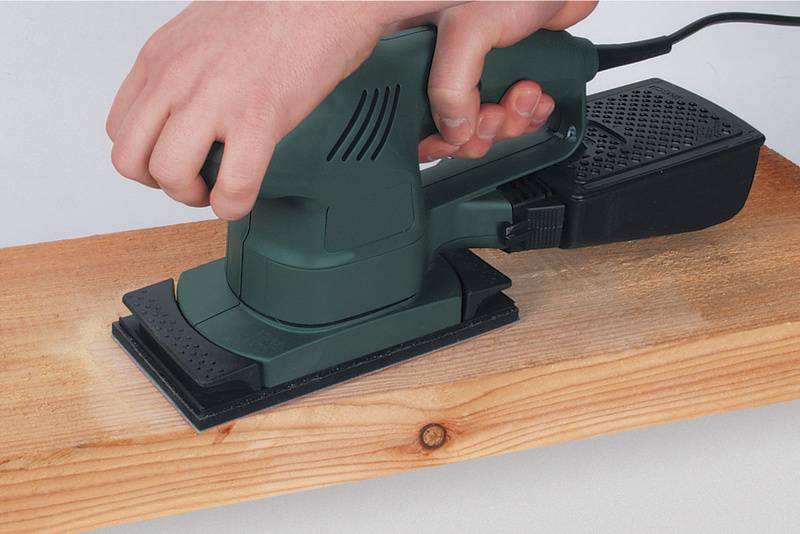 Een persoon schuurt een houten plank met een elektrische schuurmachine. Het apparaat is groen en wordt met twee handen vastgehouden.