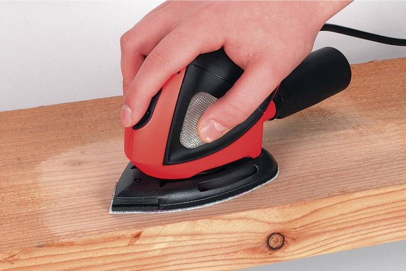 Een hand houdt een rode deltaschuurmachine vast die een houten oppervlak aan het schuren is. Het hout toont reeds bewerkte en onbewerkte gebieden.