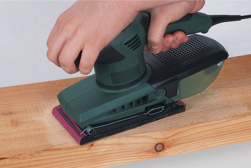 Een persoon schuurt een houten plank met een elektrische bandschuurmachine. De schuurmachine heeft een groene behuizing en ligt op een lichte plank.