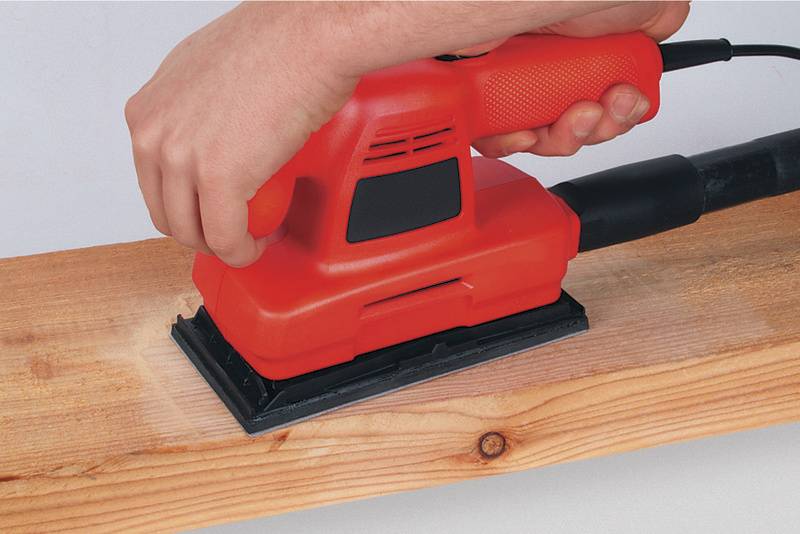 Een hand houdt een rode excentrische schuurmachine die over een houten plank wordt gevoerd om het oppervlak glad te maken.