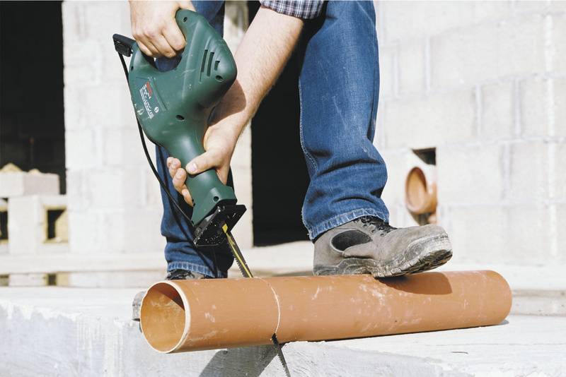 Een persoon zaagt met een elektrische decoupeerzaag een oranje kunststofbuis op een bouwplaats.