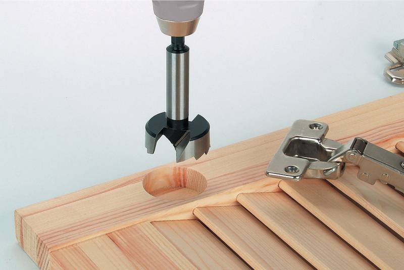 Een boormachine maakt een gat in een houten plank, naast een scharnieropening.