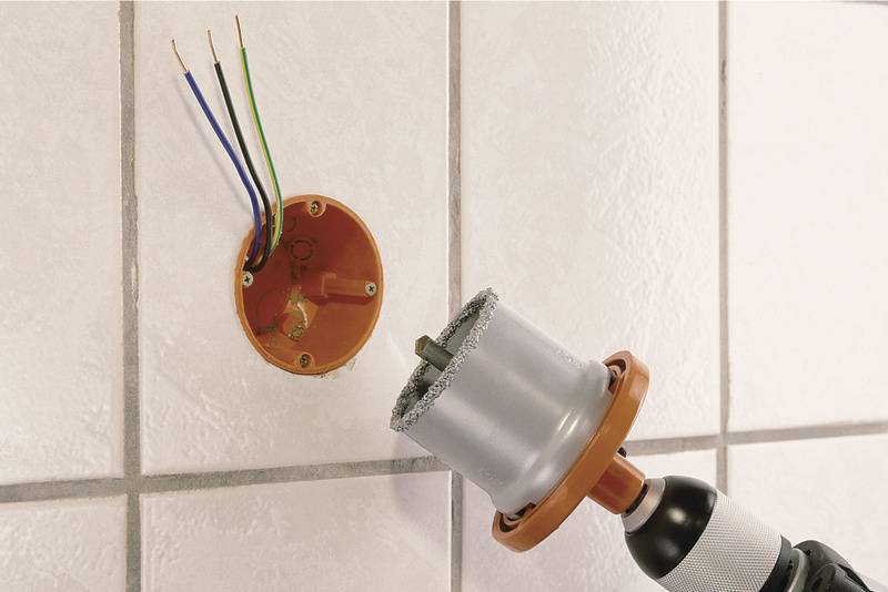 Een rond boorgat in een betegelde muur, met blootliggende kabels. Naast het gat staat een boor met een grote opzetstuk.