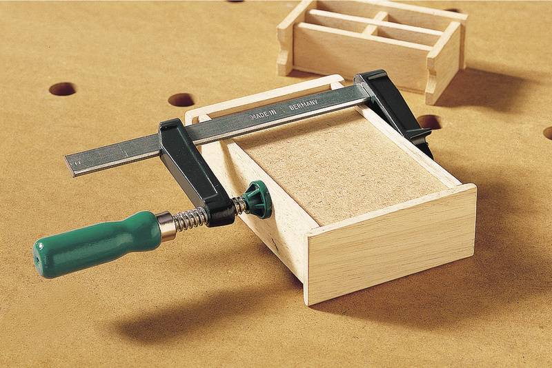 Een houten kist wordt met een klemschroef vastgezet op een werktafel. Op de achtergrond is een kleine houten doos te zien.