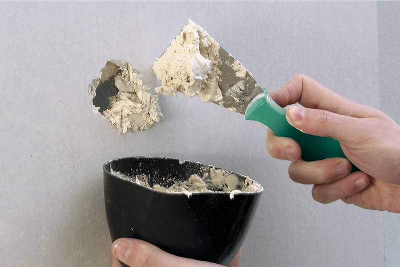 Twee handen, waarbij een hand een spatel vasthoudt met een beige-grijze massa, en de andere een zwarte container. Het gereedschap wordt beladen met plamuur.