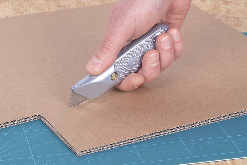 Een hand snijdt met een zilveren cuttermes precies door karton op een snijmat.