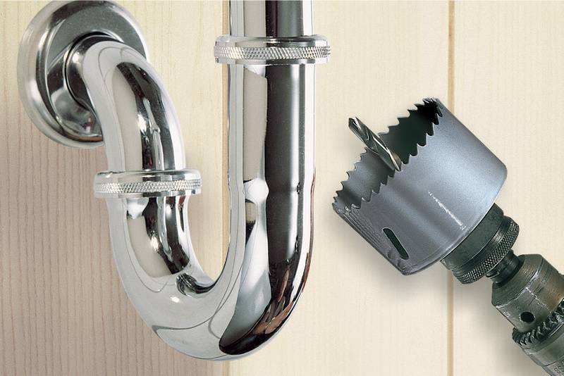 Op de afbeelding staat een verchroomd sifon-pijp voor een houten wand, naast een opzetstuk voor een boormachine.