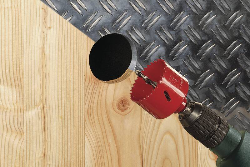 Een boor met een rode gatenzaag boort een stuk hout door, waarbij in de achtergrond metaal met een ruitpatroon zichtbaar is.
