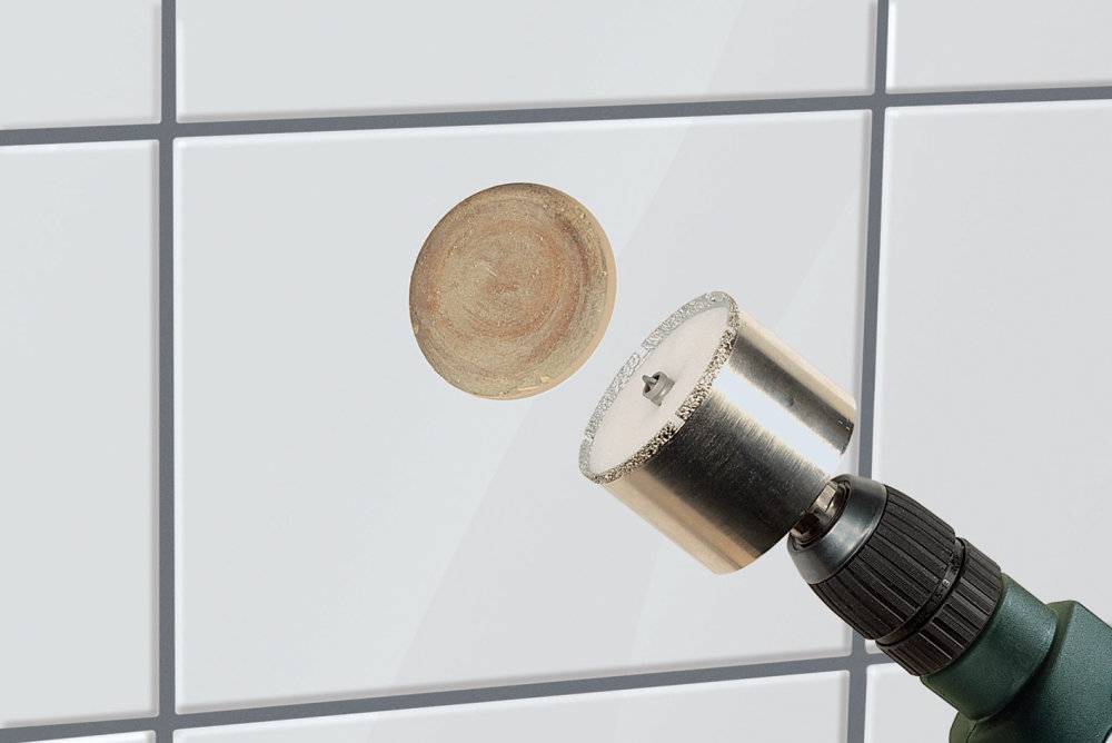 Een boormachine met gatenzaag snijdt een rond gat in een witte tegel. De boorpunt is bedekt met tegelstof.