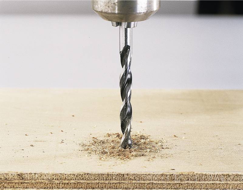 Een boormachine boort een gat in een houten plank. Houtspaanders zijn rond het gat verspreid.