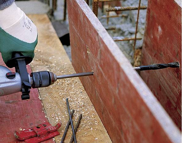 Een arbeider boort met een boormachine een gat in een houten plank op een bouwplaats. Metalen stangen en spaanders zijn zichtbaar.