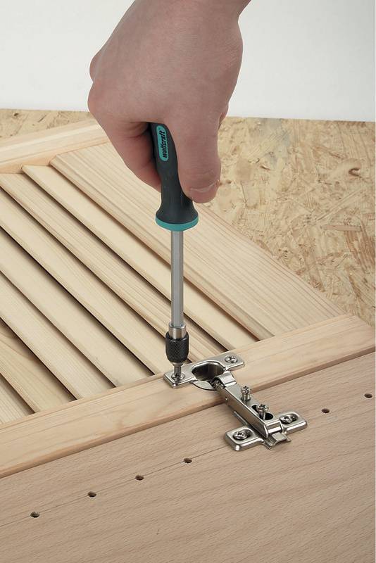 Een hand schroeft een metalen scharnier aan een houten plank met behulp van een schroevendraaier.