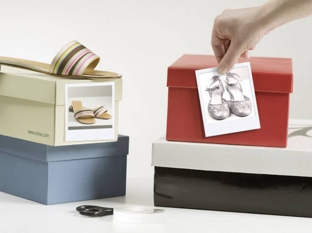 Een hand plaatst een foto van zilveren schoenen op een rode schoenendoos. Naast de foto staan andere dozen met schoenenfoto's.