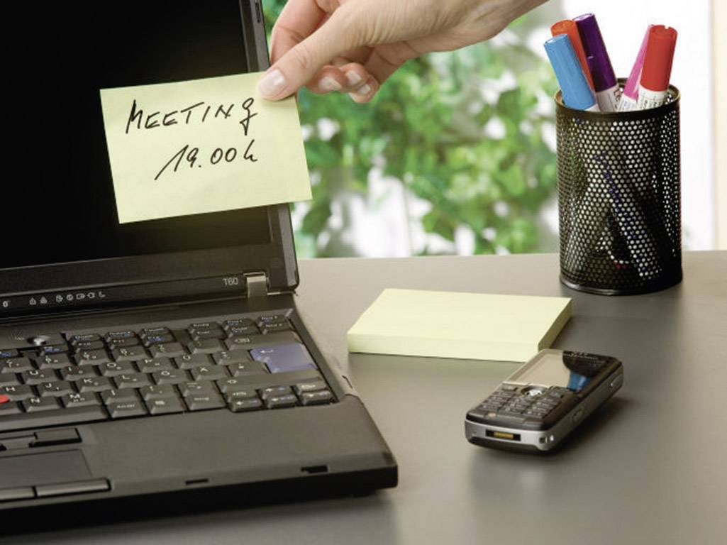 Een hand legt een notitieboekje met de tekst 'Vergadering 19:00' op een laptop. Ernaast liggen een mobiele telefoon en kleurrijke pennen.