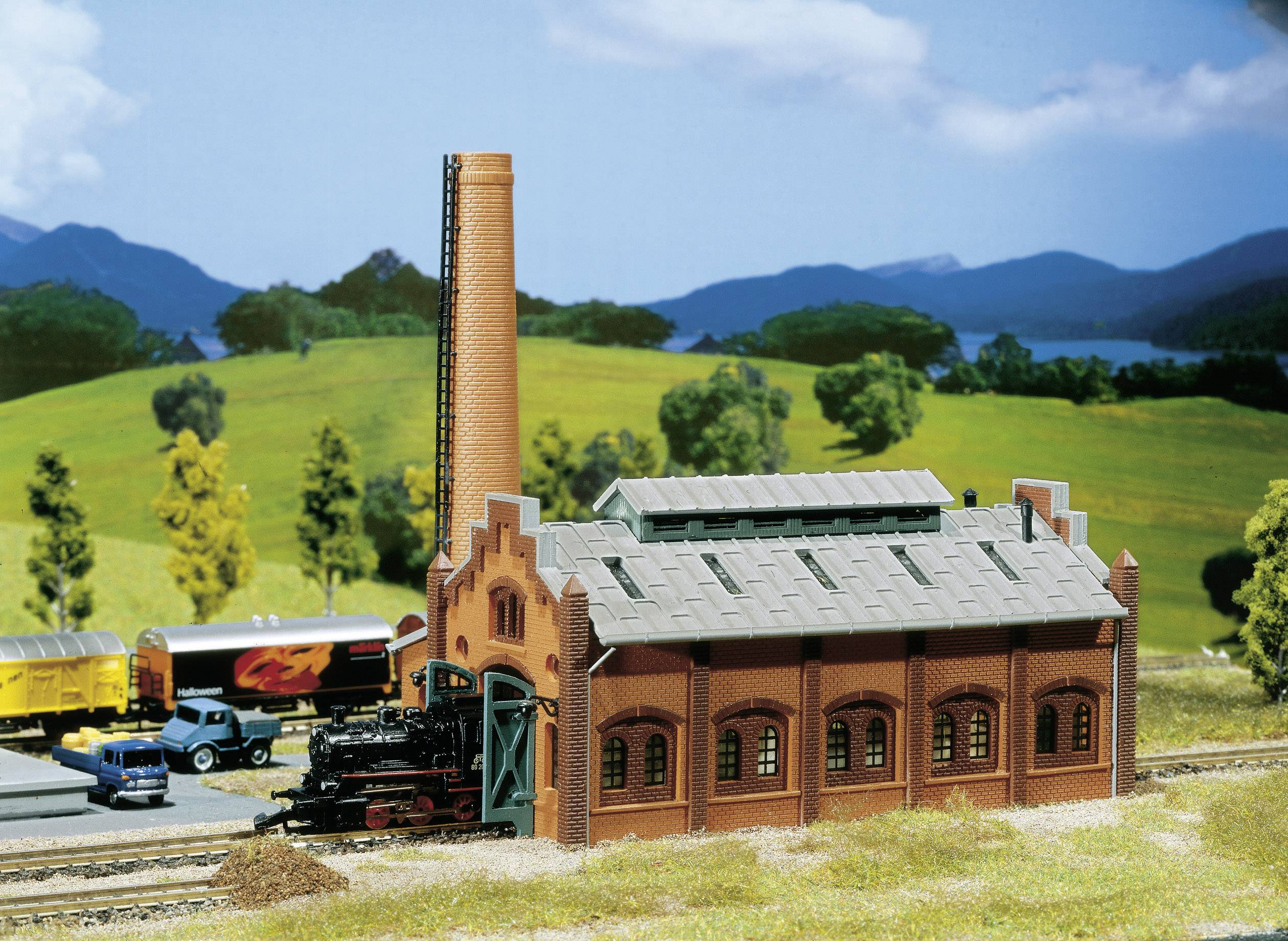Een miniatuur-stoomlocomotief staat voor een modeltreinremise in een landelijke omgeving met heuvels op de achtergrond.
