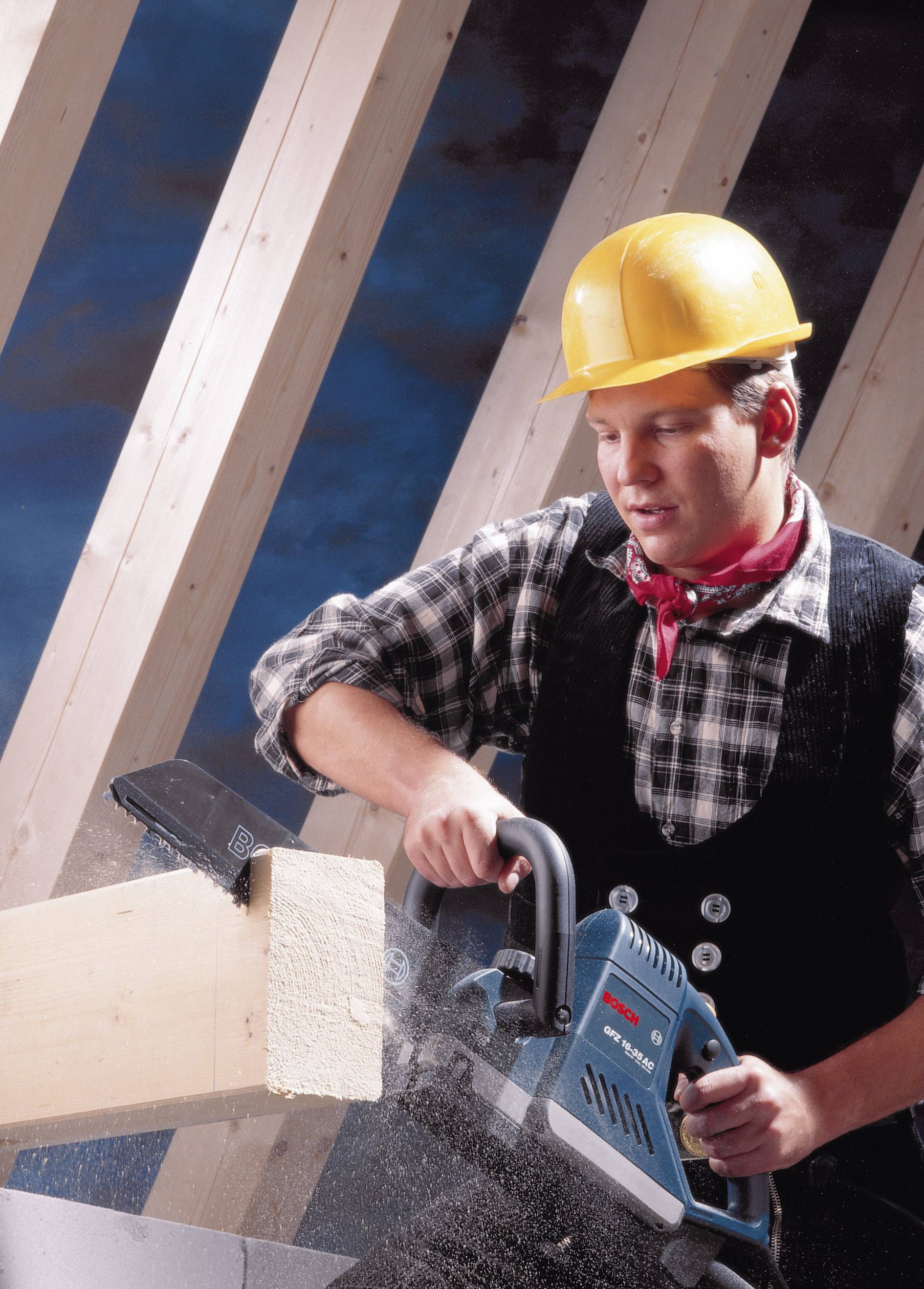 Een bouwvakker in beschermende kleding zaagt met een kettingzaag een houten plank. Op de achtergrond zijn houten balken en een blauwe lucht zichtbaar.
