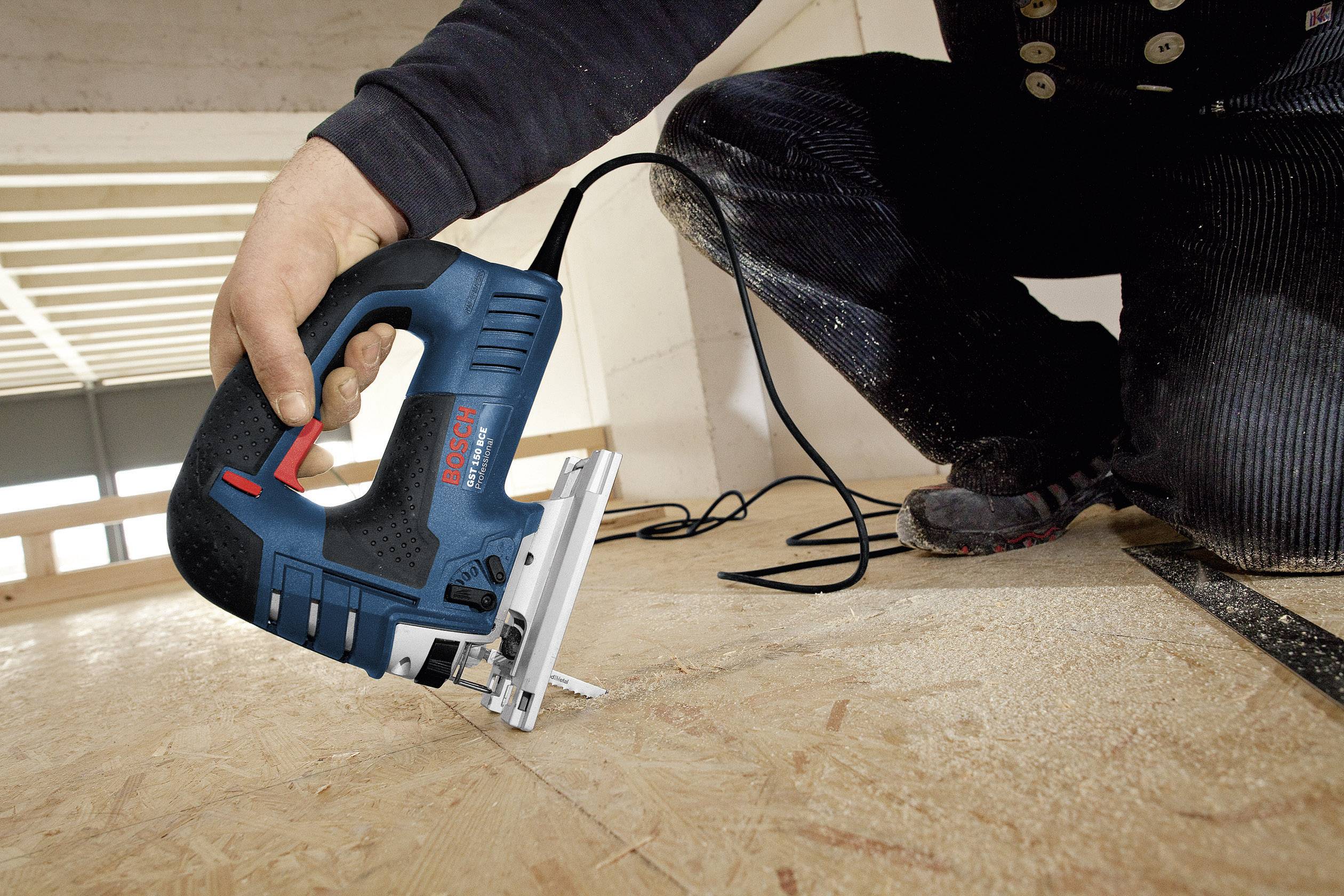 Een persoon zaagt met een blauwe decoupeerzaag een houten plank in een half afgewerkte ruimte.