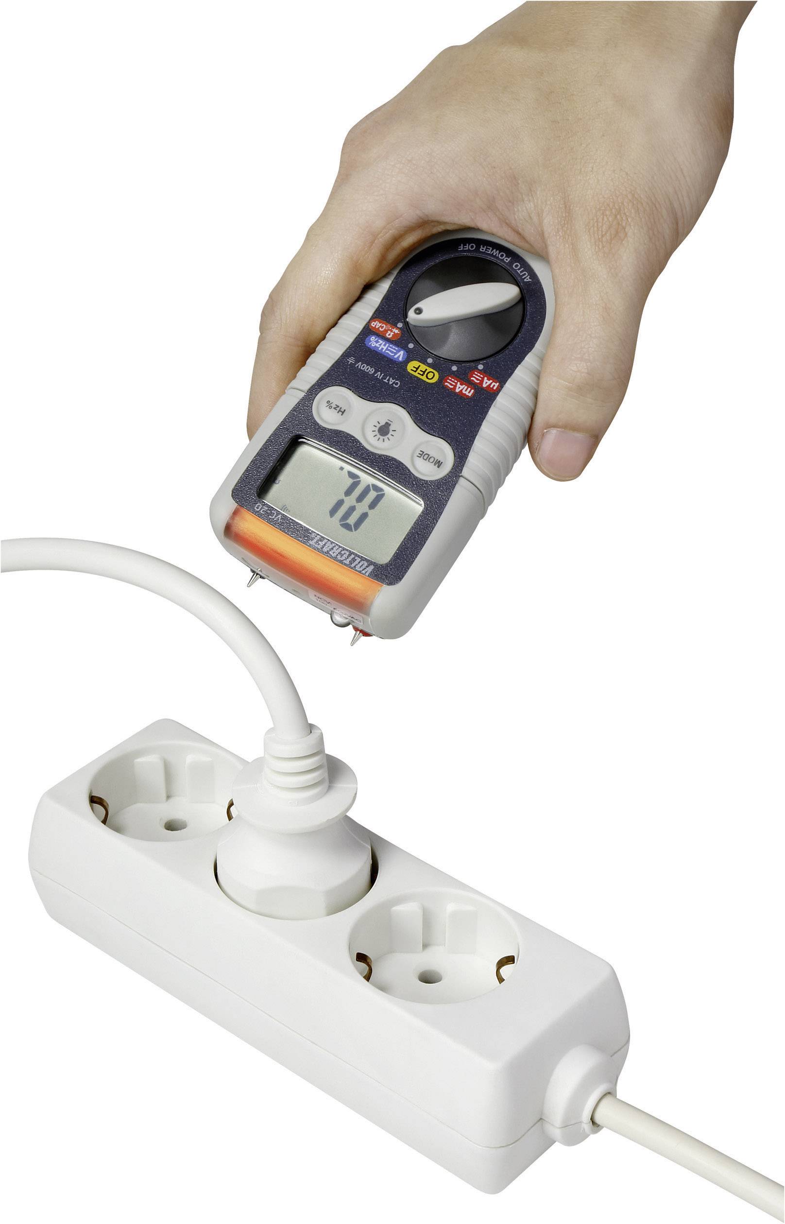 Een hand houdt een multimeter vast die de spanning van een geaard stopcontact in een stekkerdoos meet. Het display toont '70'.