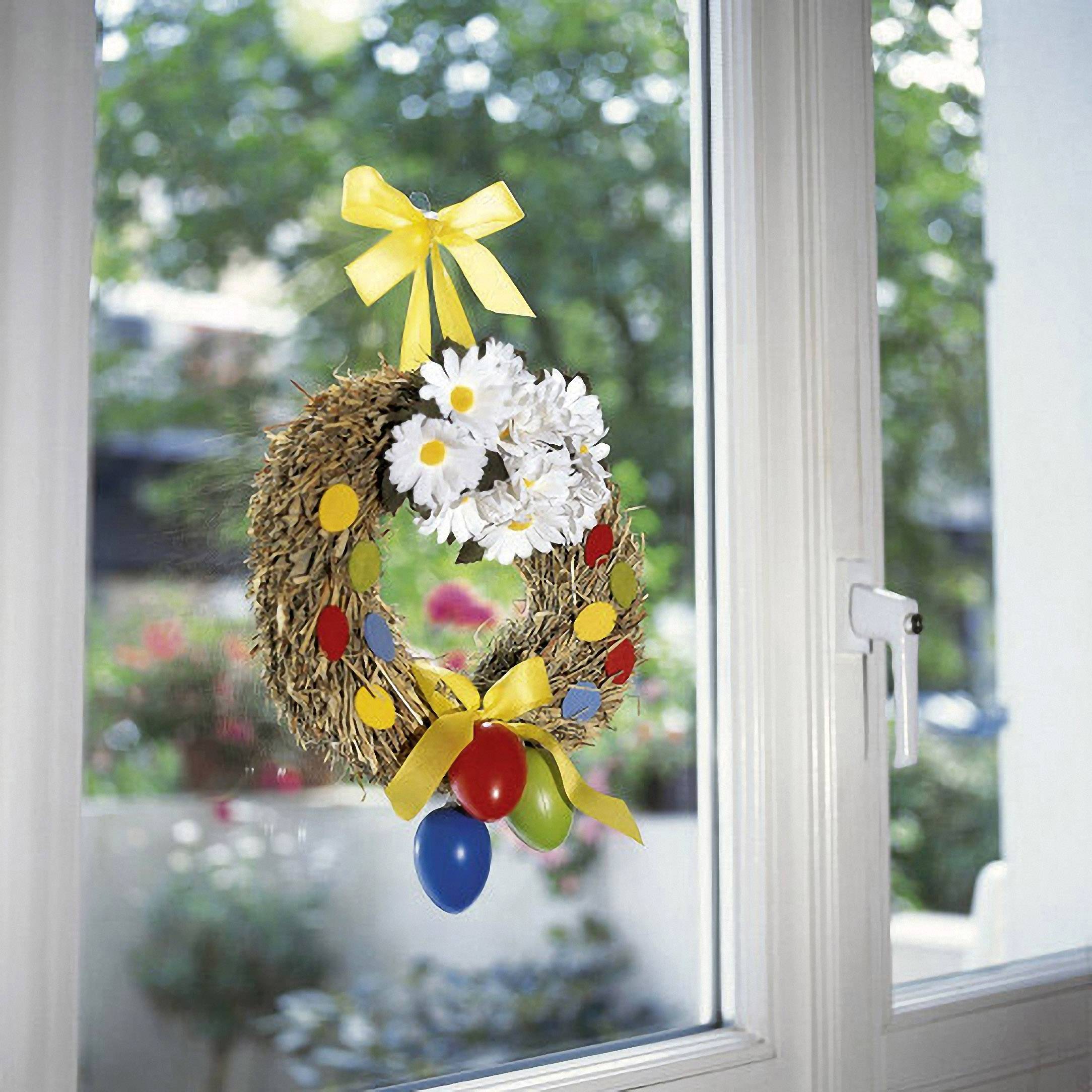 Een kleurrijke paaskrans met gele linten, witte bloemen en bonte eieren hangt voor een raam. Buiten zijn de bomen wazig te zien.
