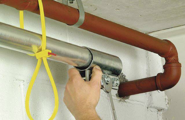Een hand bevestigt een metalen buisklem aan een buis aan het plafond. Ernaast loopt een andere rode buis parallel.