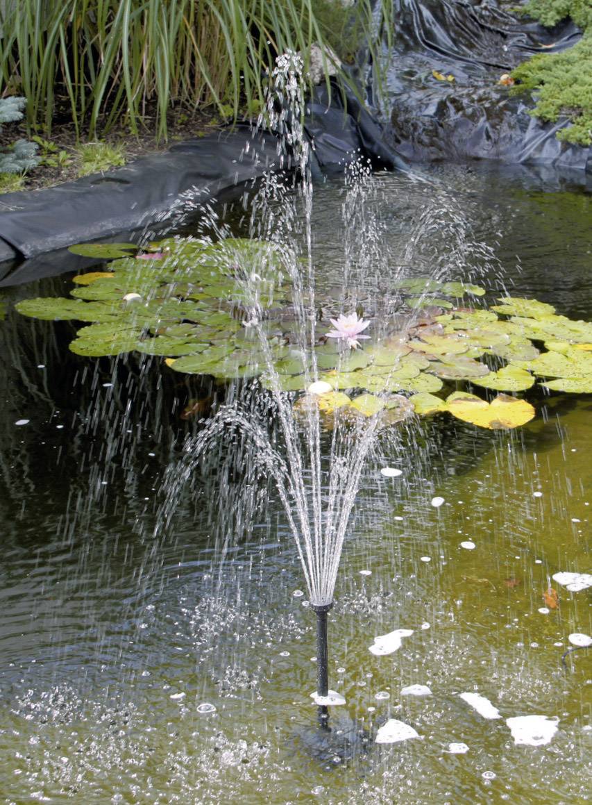 Fontein in een vijver met bladeren en een roze waterlelie op de achtergrond.