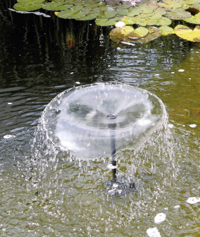 Een ronde fontein met koepelspray in een vijver, omringd door waterleliebladeren. Waterdruppels spatten in een helder patroon.