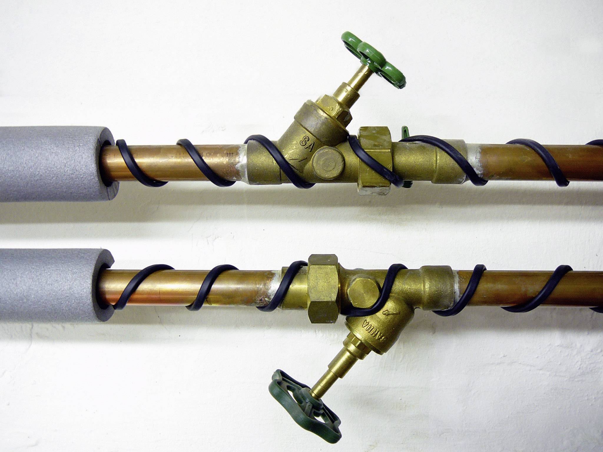 Twee geïsoleerde koperen buizen met ventielen. Elk ventiel heeft een groene handgreep. De buizen zijn gezekerd met zwarte draad.