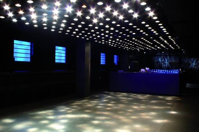 Een lege nachtclub met een verlicht plafond, blauwe lichtgevende borden aan de muren en een bar aan de rechterkant.