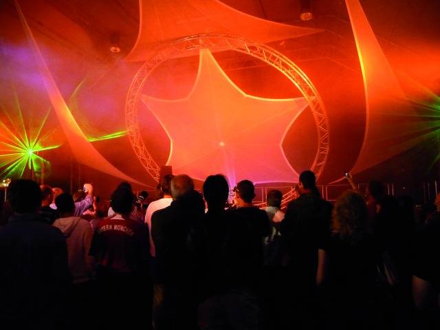 Een groep mensen danst in een club, verlicht door kleurrijke lichten en lasers, met een grote stervorming decoratie aan het plafond.