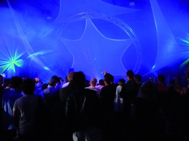 Menigte mensen voor een podium met blauw licht en lasereffecten. Toeschouwers maken foto's en genieten van de voorstelling.