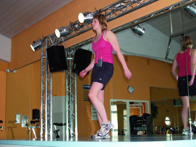 Een vrouw in een roze top en zwarte shorts doet aerobics op een podium in een fitnesscentrum, omringd door schijnwerpers en spiegels.