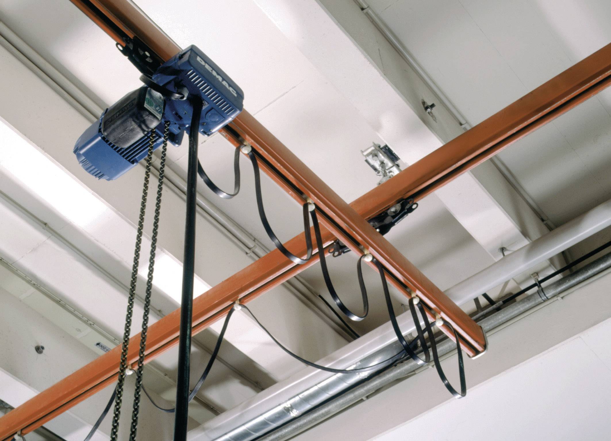 Een blauwe elektrische kettingtakel is bevestigd aan een oranje rail aan het plafond en wordt verbonden door zwarte kabels.