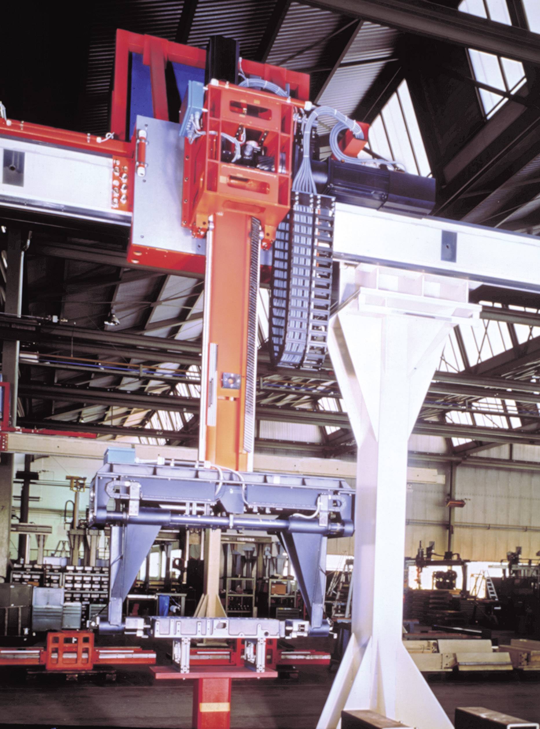 Machine in een industriehal met stalen balken en kabels. De constructie is rood, grijs en tilt onderdelen op.