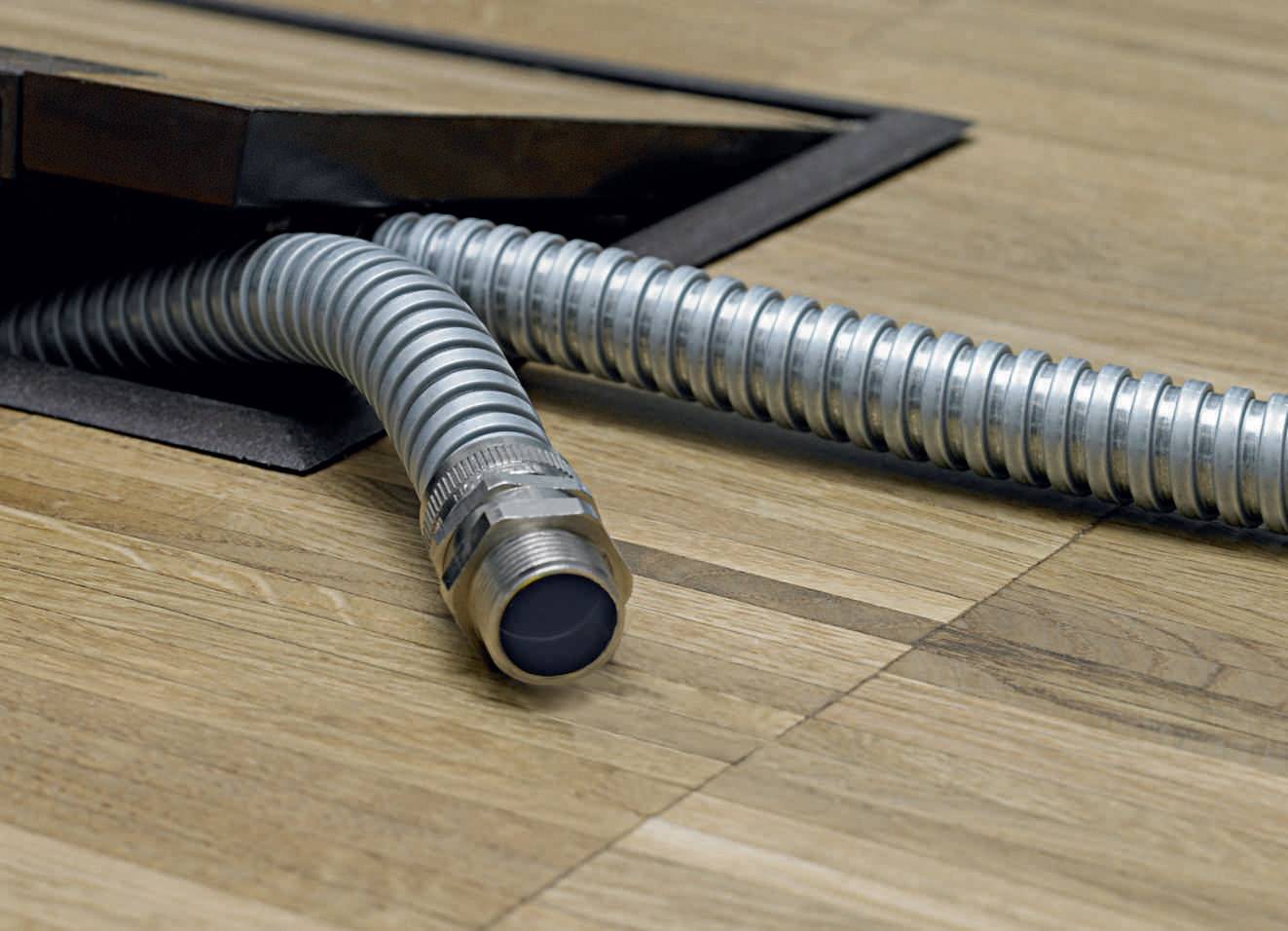 Metalen flexibele slang steekt uit een houten luik in de vloer. De scène toont een installatie of onderhoudswerk.