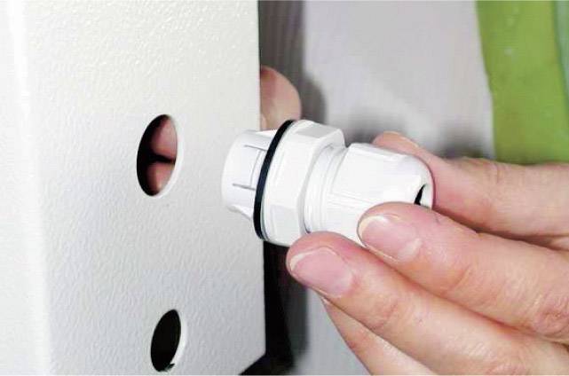 Een hand bevestigt een witte kabeldoorvoer aan een metalen behuizing met twee gaten.